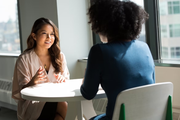 Sie sehen den Screen unseres Kurses Grundlagen der Kommunikationskompetenz. Sie sehen zwei Frauen die sich gegenüber an einem Tisch sitzen und miteinander sprechen. Die Kommunikationskompetenz ist eine Schlüsselfähigkeit für effizientes und harmonisches Zusammenarbeiten.