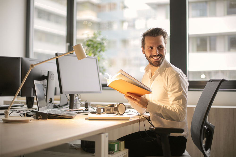 Sie sehen einen Mann an seinem Schreibtisch der seine Notizenaufschlägt und von einer anderen Person angesprochen wird. Er lächelt dabei. Es ist das Titelbild des Blogbeitrags "Digital Onboarding – Mitarbeitende erfolgreich integrieren"